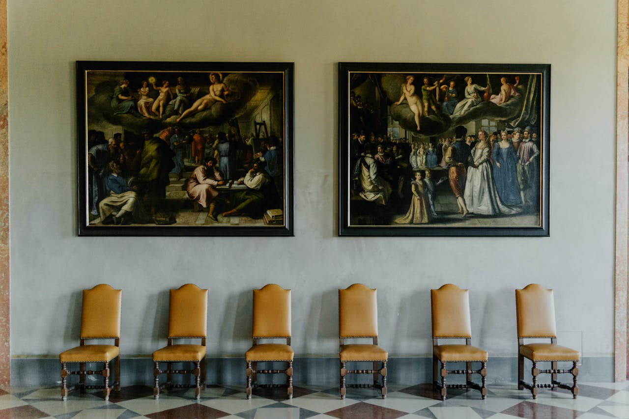 A beautifully designed interior with ornate paintings and classic wooden chairs lined up against the wall.
