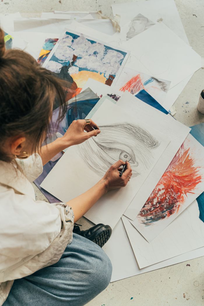 A woman sketches on the floor, surrounded by vibrant art pieces, showcasing creativity in action.