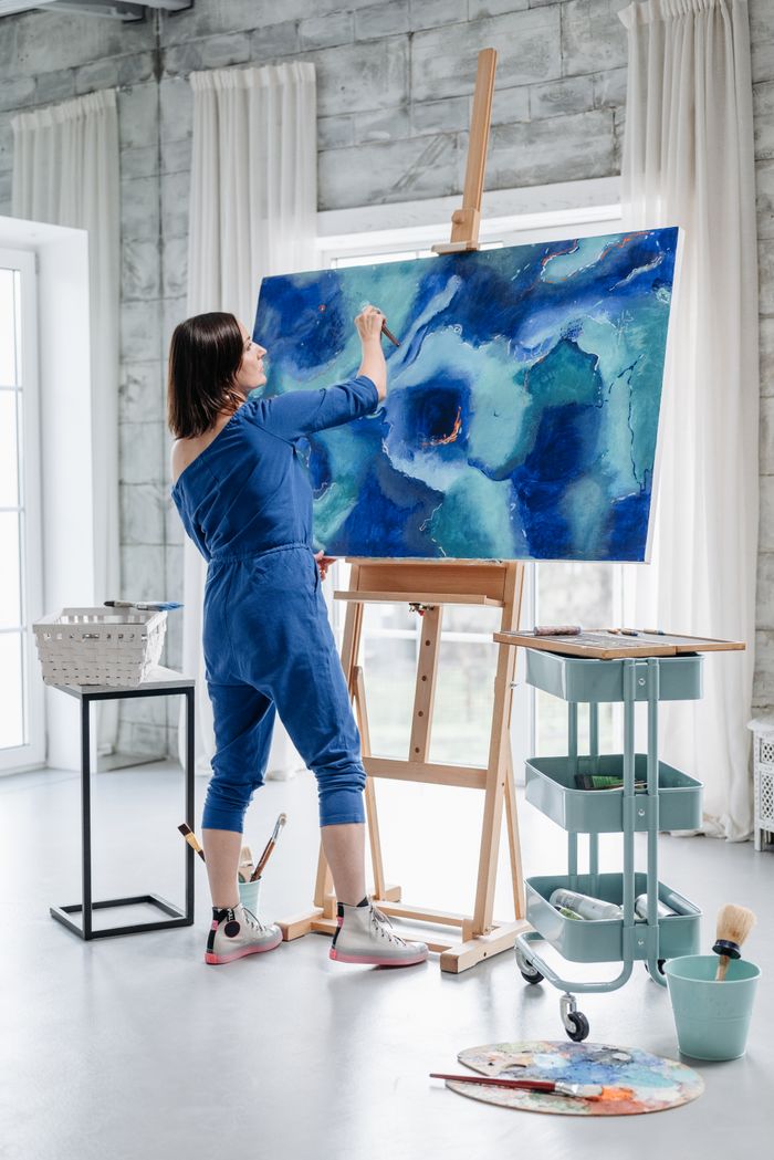 A woman artist painting a vibrant abstract blue artwork on canvas in a modern art studio.